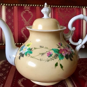 May include: Cream-colored ceramic teapot with a white handle, spout, and lid. The teapot is decorated with hand-painted floral designs in pink, blue, and green. The teapot is a vintage style and sits on a red patterned surface.