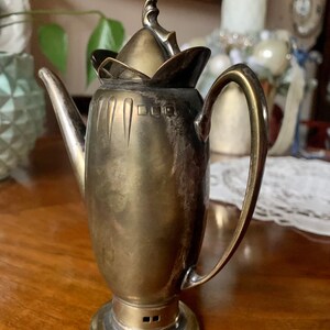 Op de afbeelding: Een vintage zilverkleurige koffiepot met een gebogen handvat en een decoratief deksel. De pot heeft een slanke, langwerpige body en staat op een ronde basis. De tuit is lang en elegant. De pot is waarschijnlijk van metaal en heeft een klassiek ontwerp.