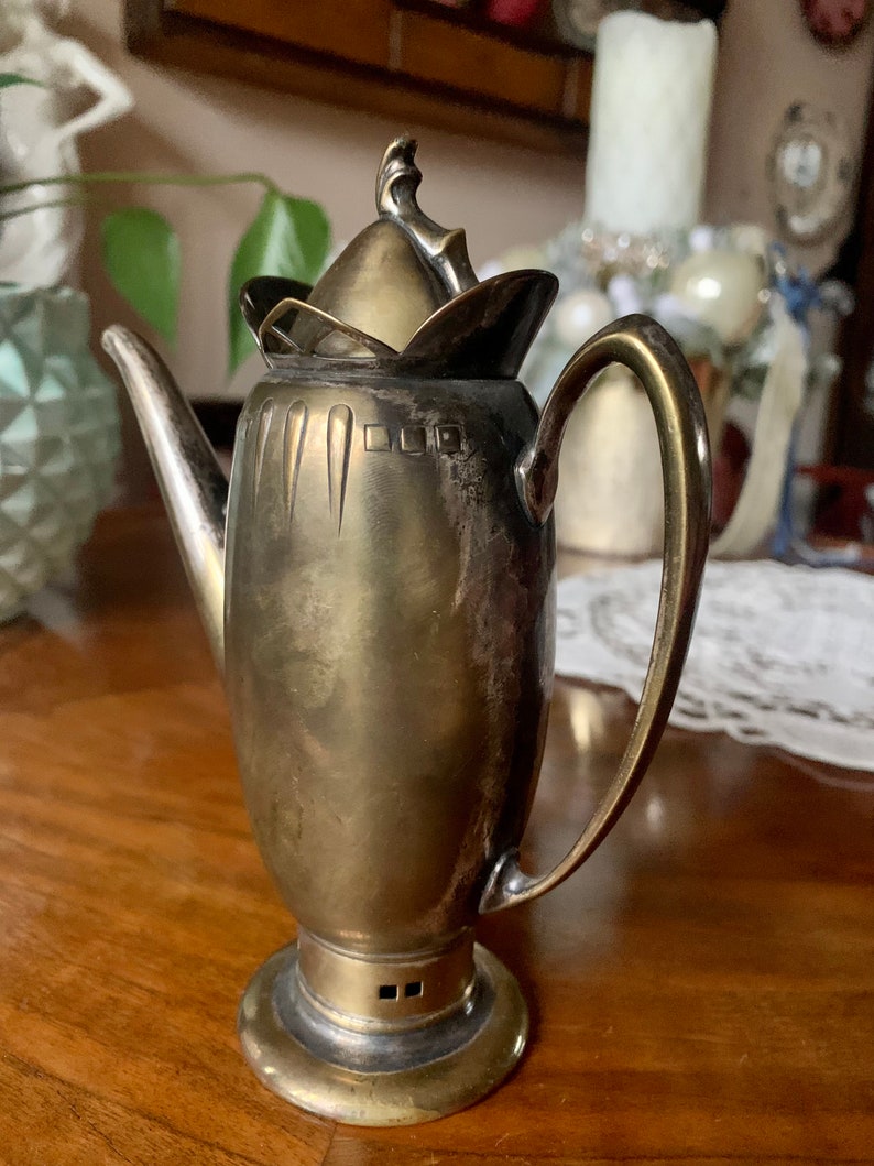 Op de afbeelding: Een vintage zilverkleurige koffiepot met een gebogen handvat en een decoratief deksel. De pot heeft een slanke, langwerpige body en staat op een ronde basis. De tuit is lang en elegant. De pot is waarschijnlijk van metaal en heeft een klassiek ontwerp.