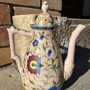 Może przedstawiać: Różowy ceramiczny imbryk z kwiatowym wzorem. Imbryk ma długi dzióbek, zakrzywiony uchwyt i pokrywkę z małym guzikiem. Kwiatowy wzór obejmuje niebieskie, czerwone, żółte i zielone kwiaty i liście. Imbryk stoi na kamiennej powierzchni.