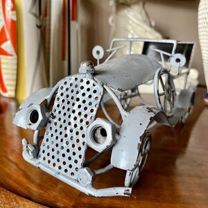 Op de afbeelding: Een grijs metalen beeld van een oldtimer. De auto is gemaakt van draad en metalen gaas en heeft een gedetailleerd rooster en koplampen. Het beeld staat op een houten tafel.