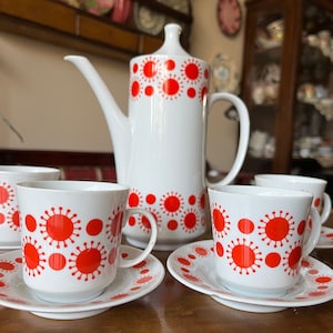 May include: A white coffee pot and four white teacups with red and orange geometric patterns. The cups and pot have a white background with red and orange circles and starbursts.