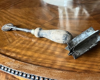 Op de afbeelding: Een vintage set van twee metalen koekjesvormen. Een vorm is een wiel met een houten handvat, de andere is een stervormige vorm.