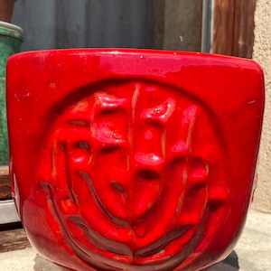 Può includere: Un vaso di terracotta rosso con un design testurizzato. Il vaso ha una forma quadrata con bordi arrotondati e un'apertura profonda e larga.