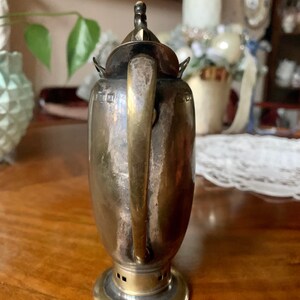 Op de afbeelding: Een vintage zilverkleurige theepot met een messing handvat en voet. De theepot heeft een gebogen handvat en een kleine decoratieve knop op het deksel. De body van de theepot heeft een cilindrische vorm met een licht uitlopende voet.