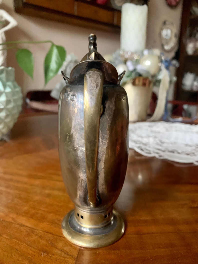 Op de afbeelding: Een vintage zilverkleurige theepot met een messing handvat en voet. De theepot heeft een gebogen handvat en een kleine decoratieve knop op het deksel. De body van de theepot heeft een cilindrische vorm met een licht uitlopende voet.