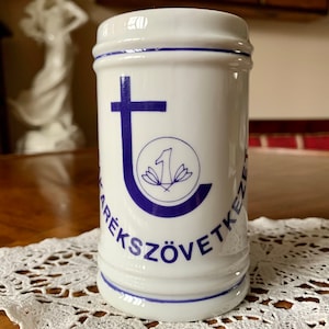 May include: White ceramic jar with blue accents and text. The jar features a blue cross and a circular emblem with a number one and floral design. The text "TÁRÉKSZÖVETKEZET" is printed around the jar.