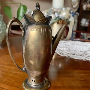 Op de afbeelding: Een vintage zilverkleurige metalen theepot met een gebogen handvat en een lange tuit. De theepot heeft een decoratief deksel en een ronde basis. Het metaal heeft een aangetaste uitstraling, wat op leeftijd duidt. De theepot wordt waarschijnlijk gebruikt voor het serveren van thee.