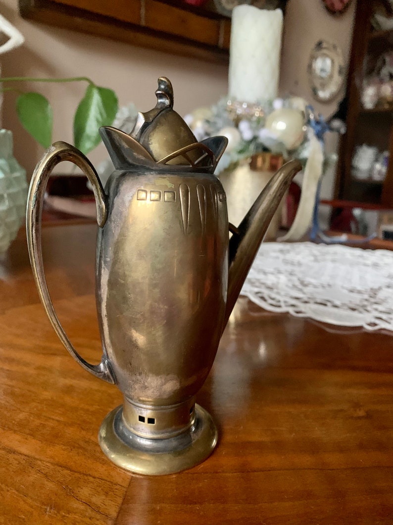 Op de afbeelding: Een vintage zilverkleurige metalen theepot met een gebogen handvat en een lange tuit. De theepot heeft een decoratief deksel en een ronde basis. Het metaal heeft een aangetaste uitstraling, wat op leeftijd duidt. De theepot wordt waarschijnlijk gebruikt voor het serveren van thee.