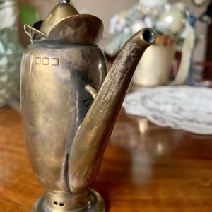 Op de afbeelding: Een vintage zilveren en messing theepot met een gebogen tuit en een klein deksel. De theepot heeft een ronde basis en een handvat bovenop het deksel. Het metaal heeft een aangetaste uitstraling, wat op leeftijd duidt. De theepot staat op een houten oppervlak.