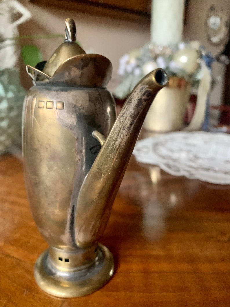 Op de afbeelding: Een vintage zilveren en messing theepot met een gebogen tuit en een klein deksel. De theepot heeft een ronde basis en een handvat bovenop het deksel. Het metaal heeft een aangetaste uitstraling, wat op leeftijd duidt. De theepot staat op een houten oppervlak.