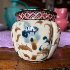 Può includere: Un piccolo vaso in ceramica bianca con un bordo rosa e oro. Il vaso è decorato con un motivo floreale in tonalità di blu, verde e marrone.