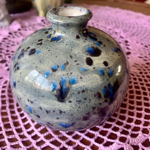 Può includere: Un piccolo vaso rotondo in ceramica blu e bianca con una glassa macchiettata. Il vaso ha un'ampia apertura in alto e un collo stretto.