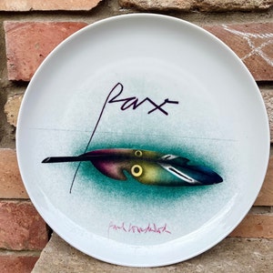 May include: White ceramic plate with a teal green background and a colorful feather design. The plate is signed "Pax" and "Paul Wunderlich".