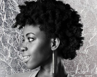 May include: Black and white portrait of a person with short, dark, curly hair and a silver earring. The person is looking to the side with a slight smile. The background is a textured silver surface.