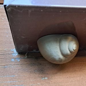 Op de afbeelding: Een bruine metalen doos met een lichtgekleurde, schelpvormige voet. De doos staat op een houten oppervlak. De schelpvoet is een decoratief element dat een unieke touch toevoegt aan het ontwerp van de doos.