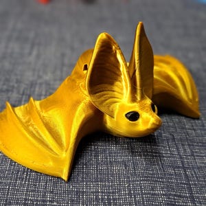 Könnte beinhalten: Eine metallisch goldfarbene Fledermausfigur mit großen, spitzen Ohren und schwarzen Augen. Die Fledermaus ist in einer ruhenden Position mit teilweise ausgebreiteten Flügeln dargestellt, die eine glatte, reflektierende Oberfläche zeigen.