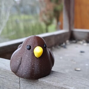 May include: A small, rounded, dark brown bird figurine with a smooth, slightly speckled texture. It features two small, round black eyes and a bright yellow, triangular beak. The figurine is positioned on the edge of a wooden structure.