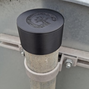 Peut inclure: Un objet cylindrique noir avec une surface texturée et un emblème détaillé sur le dessus. L'objet est monté sur un poteau métallique, suggérant un panneau ou un marqueur. L'emblème semble être un motif stylisé.