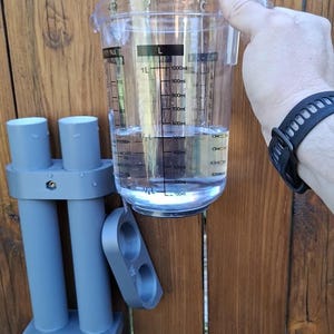 May include: A clear plastic measuring pitcher with black markings up to 1000ml, held next to a grey dual-tube wall mount bracket. The pitcher contains liquid filled to the 400ml line, and the bracket features two cylindrical holders.