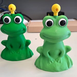 Può includere: Due statuette di rana verdi con grandi occhi neri e bianchi e un'antenna gialla. Le rane sono sedute su una base verde. Lo sfondo è una superficie bianca con componenti elettronici e una scatola di cartone.
