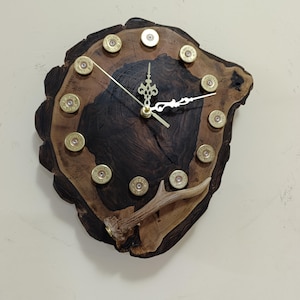 May include: A rustic wall clock crafted from a dark wood slab with a natural edge. The clock face features brass bullet casings as hour markers and delicate gold-colored hands. A small antler adds a unique decorative element.