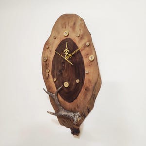 May include: A rustic wooden wall clock with a natural edge. The clock face is a dark brown wood grain, surrounded by lighter wood. Gold-colored hands and hour markers are present. A deer antler is attached to the bottom. The clock measures approximately 20 inches in height.