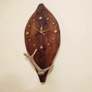 May include: A rustic wall clock with a dark brown, leaf-shaped wooden face. The clock features gold-colored hands and hour markers, and is accented with a deer antler. The clock measures approximately 24 inches in height.
