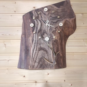 May include: A rustic wall clock crafted from a live edge wood slab. The clock features a natural wood grain pattern, metallic accents, and a decorative antler element. The clock hands are silver. The clock is mounted on a wooden wall.