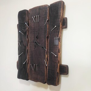 May include: A rustic wooden wall clock with a dark brown finish. The clock face is made of three vertical wooden planks with silver nail accents marking the hours. The clock hands are black.