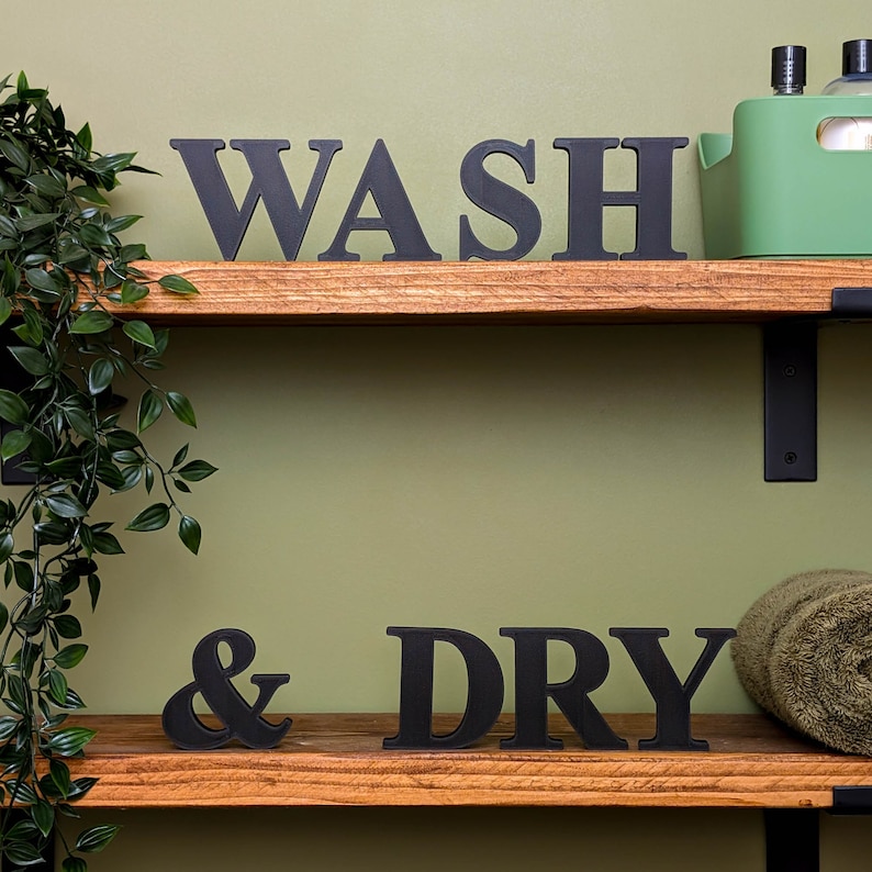 May include: Two wooden shelves with black metal brackets. The top shelf has the word "WASH" in black block letters. The bottom shelf has the word "& DRY" in black block letters.