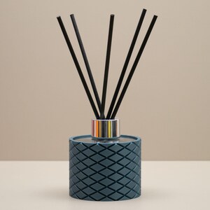 May include: A teal reed diffuser with a diamond pattern. It has a silver and gold collar and black reeds. The background is a neutral beige. The diffuser is designed to release fragrance into a room.