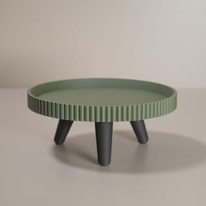 May include: A sage green, round decorative tray with a ridged edge, supported by three black legs. The tray is approximately 30 cm in diameter, suitable for displaying small items.