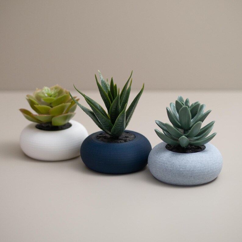 May include: Three small artificial succulent plants in round, textured pots. The succulents are green, with one in a white pot, one in a navy blue pot, and one in a light gray pot. The pots are arranged in a row.
