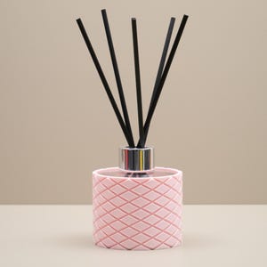 May include: A pink reed diffuser with a geometric diamond pattern. The diffuser has a silver collar and black reeds. The background is a neutral beige colour. This decorative item is designed for home fragrance.