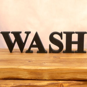 May include: Black wooden letters spelling out the word "WASH" on a wooden surface.