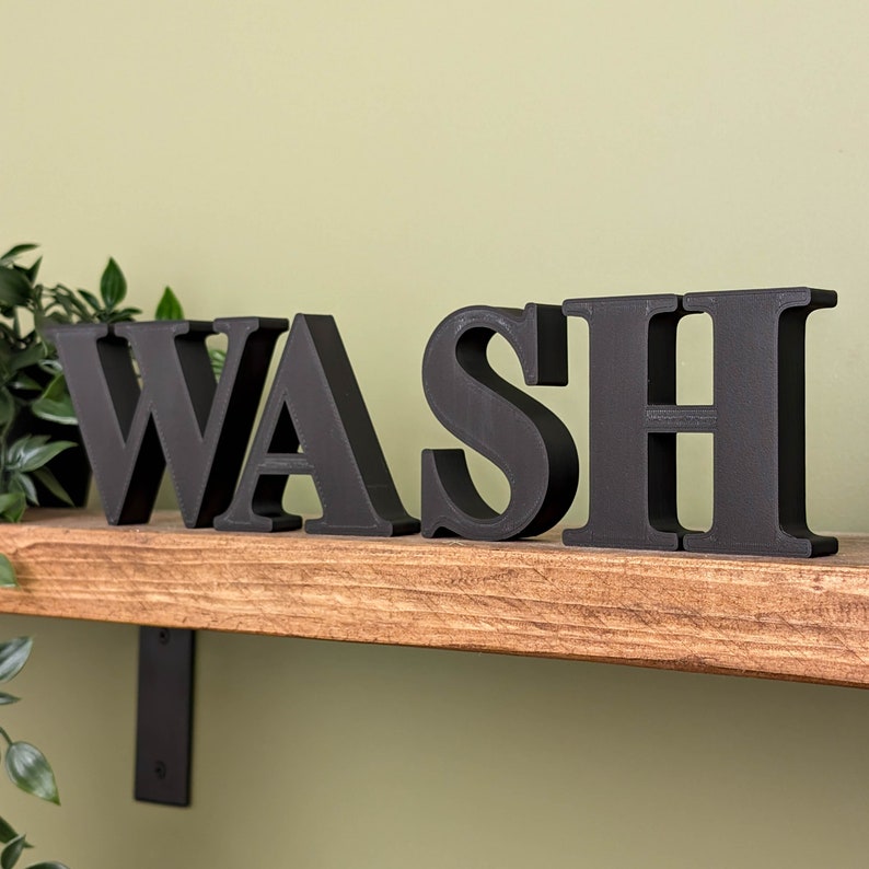 May include: Black wooden letters spelling out the word "WASH" are displayed on a wooden shelf. The letters are freestanding and have a slightly textured finish.