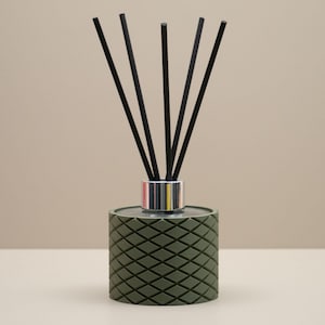 May include: A green diffuser with a diamond pattern, silver collar, and black reeds. The fragrance diffuser sits on a white surface with a neutral background. The reeds are arranged in a fan shape, adding a decorative element.