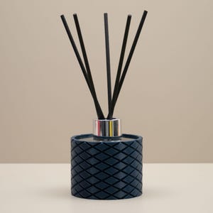 May include: A dark blue reed diffuser with a diamond pattern. The diffuser has a silver collar and contains several black reeds. The product is designed to release fragrance into a room. The background is a neutral beige.