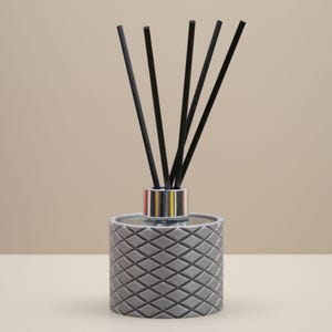 May include: A grey reed diffuser with a diamond pattern on the container. The diffuser has a silver collar and black reeds. The background is a neutral beige colour. This home fragrance item is designed to release scent into the air.