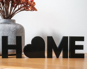 May include: Black wooden letters spelling "HOME" with a black heart in between the "H" and "O". The letters are sitting on a wooden surface.