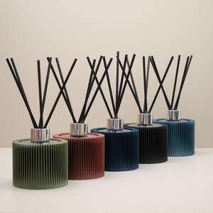 May include: Five reed diffusers in a row, each with a different coloured base: green, red, navy, black, and teal. Each diffuser has a silver collar and a bundle of black reeds. The bases have a ribbed, cylindrical design.