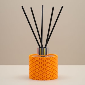 May include: An orange reed diffuser with a diamond pattern. The diffuser has a silver collar and contains black reeds. The product is designed to release fragrance into a room. The background is a neutral beige.