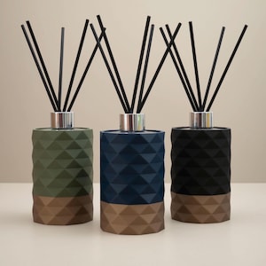 May include: Three reed diffusers with black reeds. The cylindrical bottles feature a geometric pattern in olive green, navy blue, and black, with a silver collar and a brown base. These diffusers are designed for home fragrance, a stylish addition.