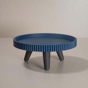 May include: A navy blue, round decorative tray with a ridged edge and three black legs. The tray is approximately 30 cm in diameter and stands about 15 cm tall. The design is modern and minimalist.