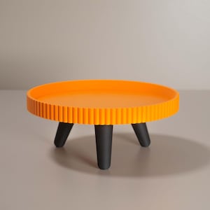May include: A small, round orange table with a ridged edge and three black legs. The table is a vibrant shade of orange and is approximately 30 cm in diameter.