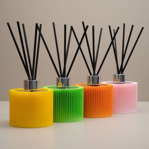 May include: Four brightly coloured reed diffusers in yellow, green, orange, and pink. Each has a silver collar and black reeds. The cylindrical bases have a ribbed texture, creating a modern look for home fragrance.