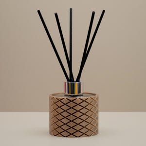 May include: A brown reed diffuser with a geometric diamond pattern. The diffuser has a silver collar and contains several black reeds. The background is a neutral beige colour, and the diffuser sits on a white surface.