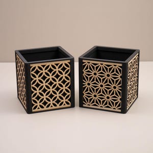 May include: Two square wooden boxes with black frames and geometric cutouts. One box features a pattern of interlocking circles, the other a star design. The interior of the boxes is black.