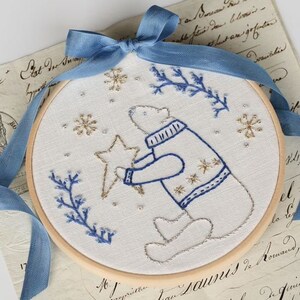 May include: Embroidered hoop art featuring a white polar bear holding a gold star, surrounded by blue and gold snowflakes and blue sprigs on a white background.